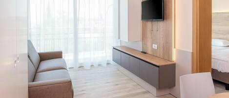A modern hotel room with a sofa, a small table, a TV mounted on a wooden panel, and a bed with a headboard.