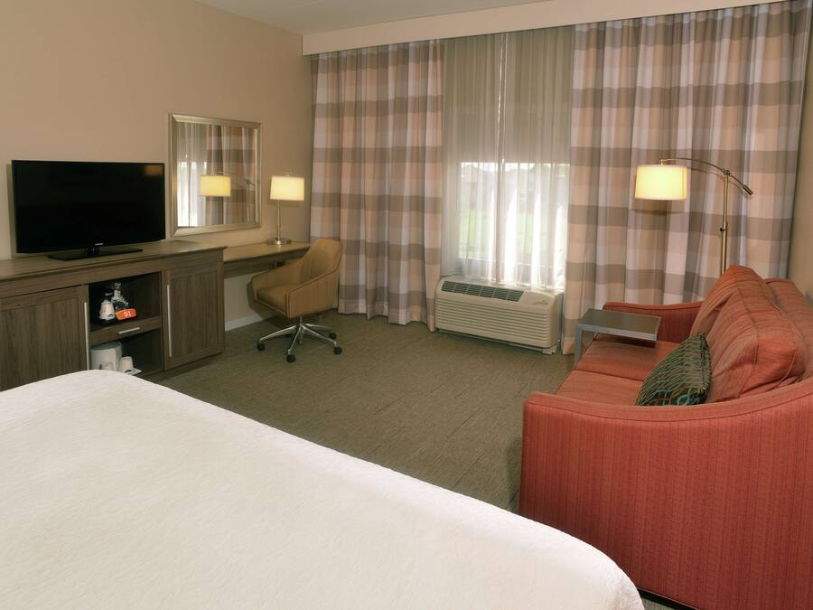 Habitación de hotel con cama, escritorio con televisor, una silla, un sofá y una ventana con cortinas.