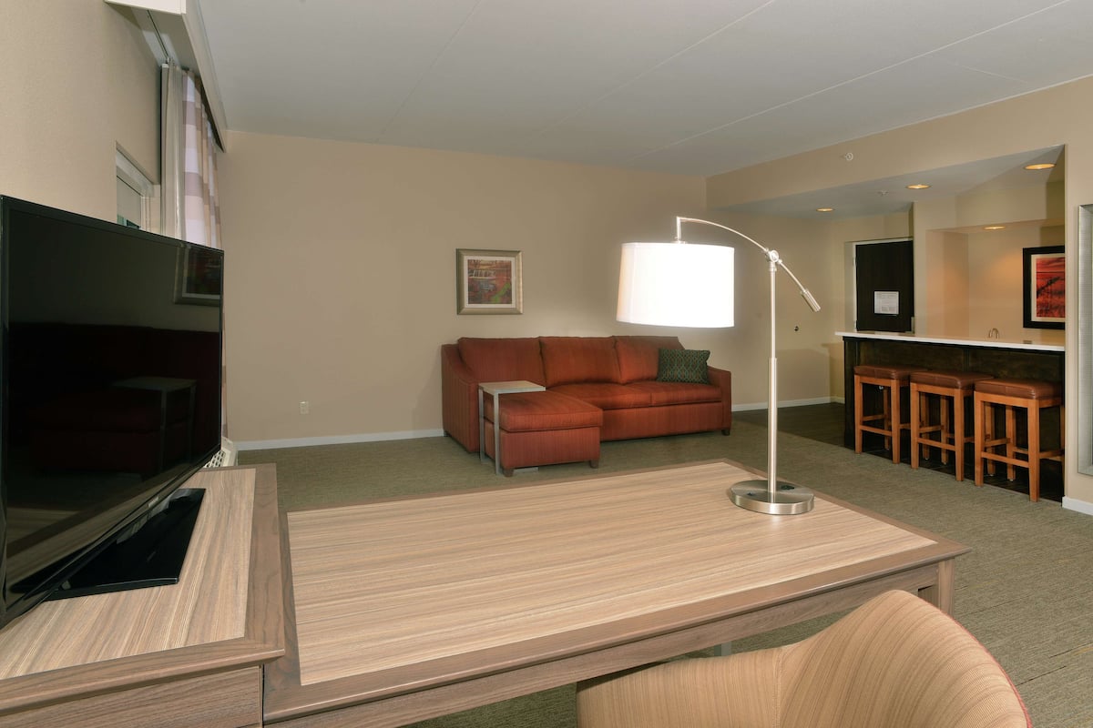 A modern living room with a flat-screen TV, a wooden desk, a sofa, and a bar area with stools.