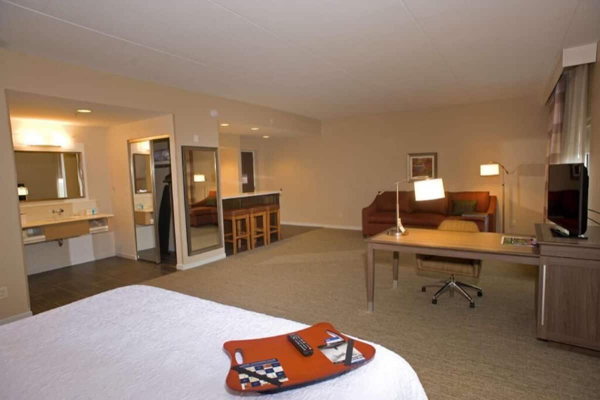 A hotel room with a bed, a desk, a chair, a TV, and a bathroom with a mirror and sink.