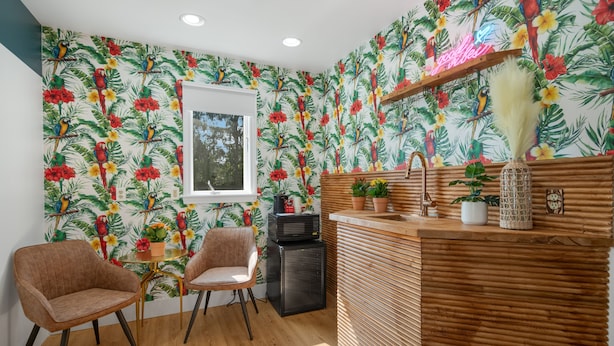 A kitchen with a tropical wallpaper, wooden counter, two chairs, and a small refrigerator.