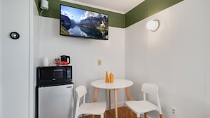 A compact kitchen with a microwave, coffee maker, and a small refrigerator. A round table with two white chairs is positioned in front of a flat-screen TV mounted on the wall, displaying a scenic landscape.