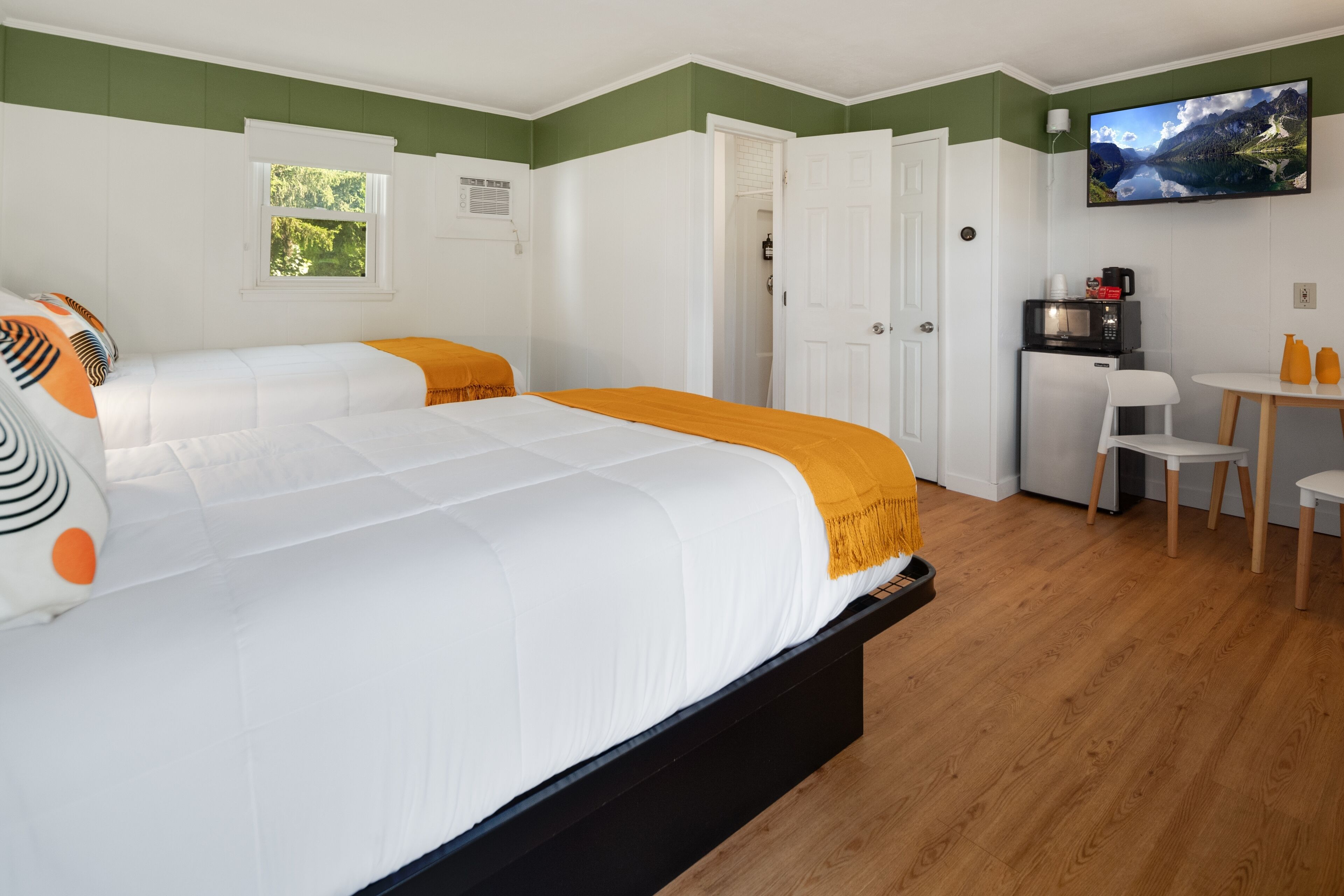 A hotel room with two beds, a TV, a desk with a chair, and a small table with orange bottles.
