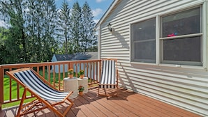 A wooden deck with two striped lounge chairs and a small table, overlooking a green lawn and trees.