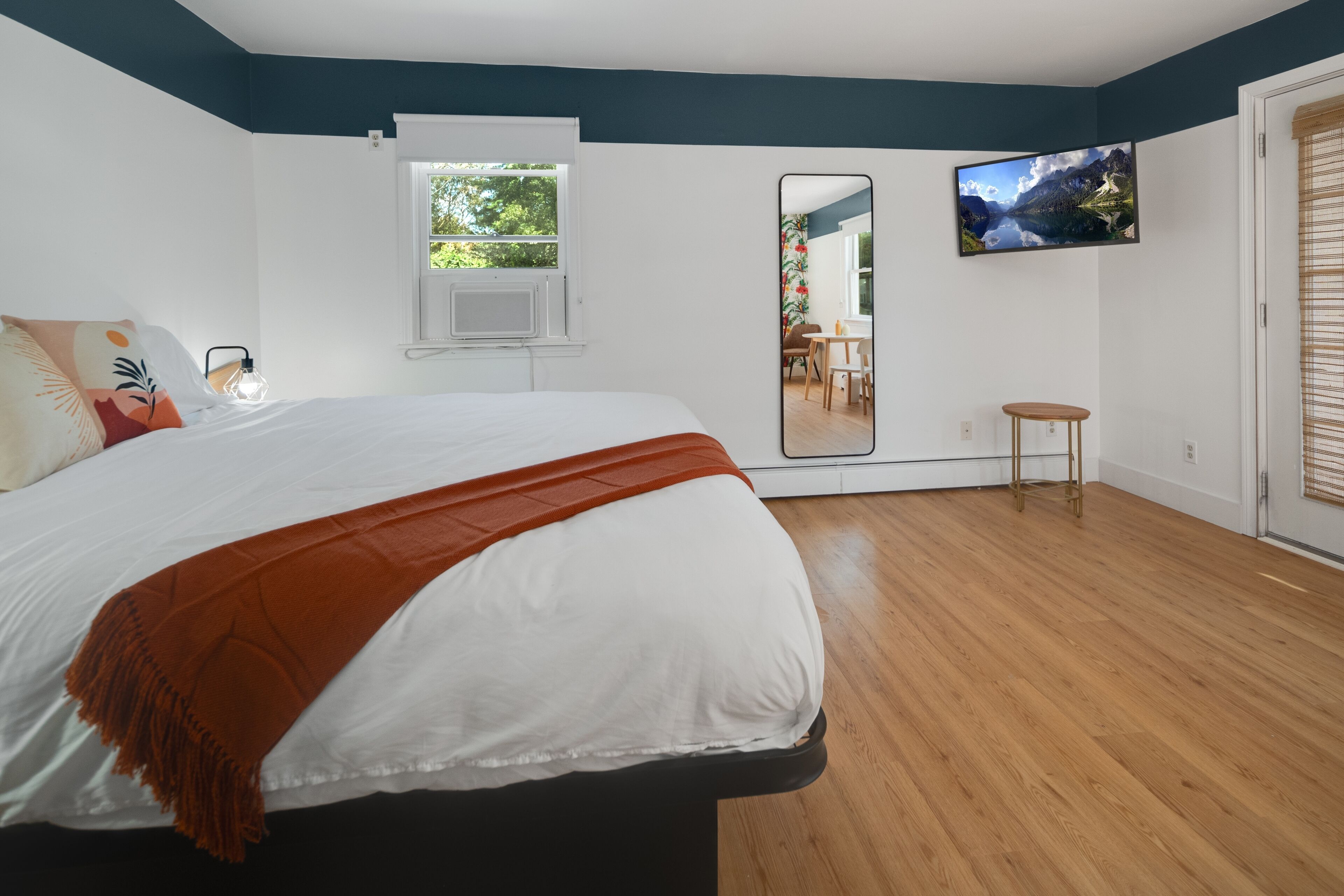 A bedroom with a large bed, a window, a mirror, and a television on the wall.