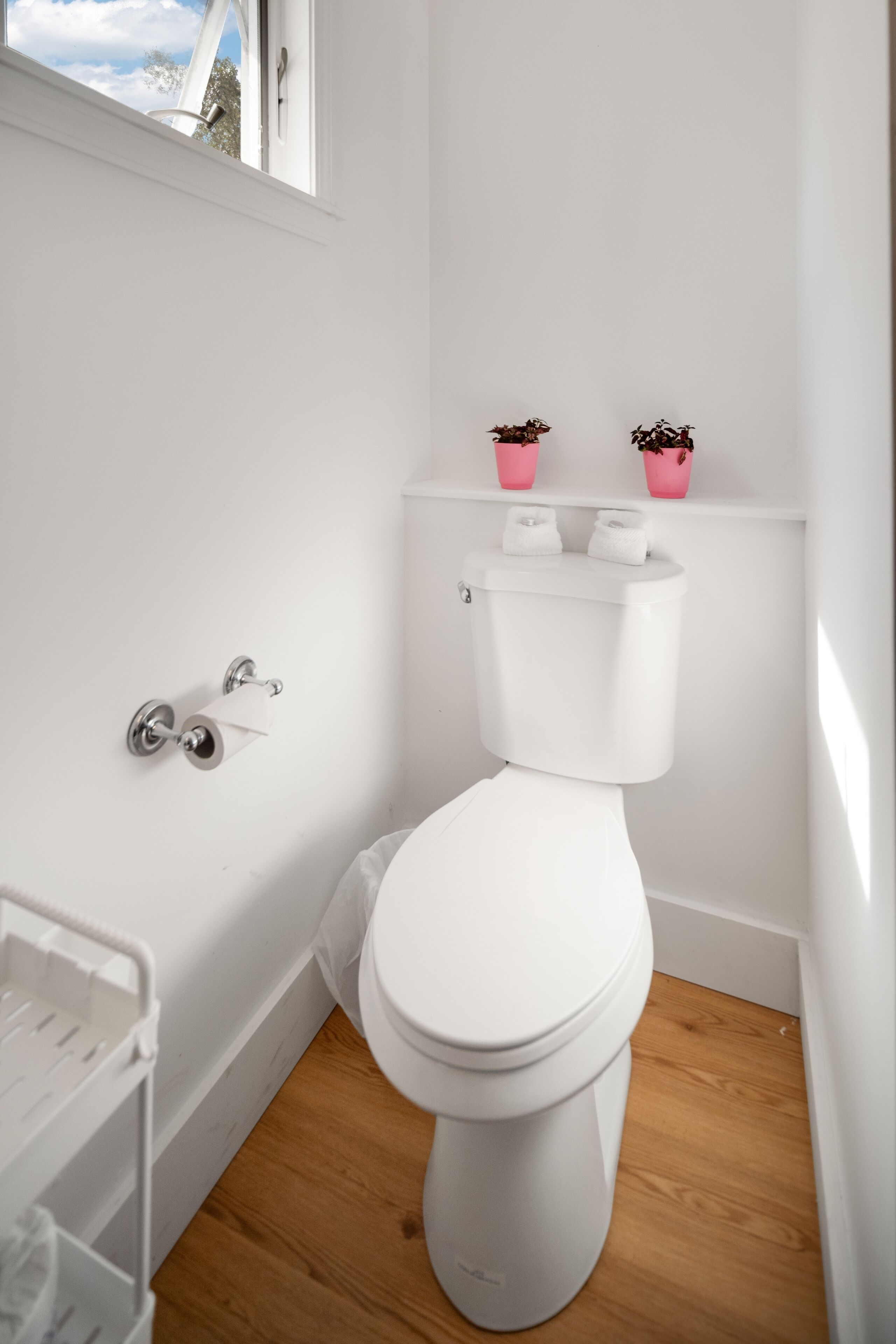 A white toilet with a white tank, a toilet paper holder, and two pink potted plants on a white shelf above the toilet.