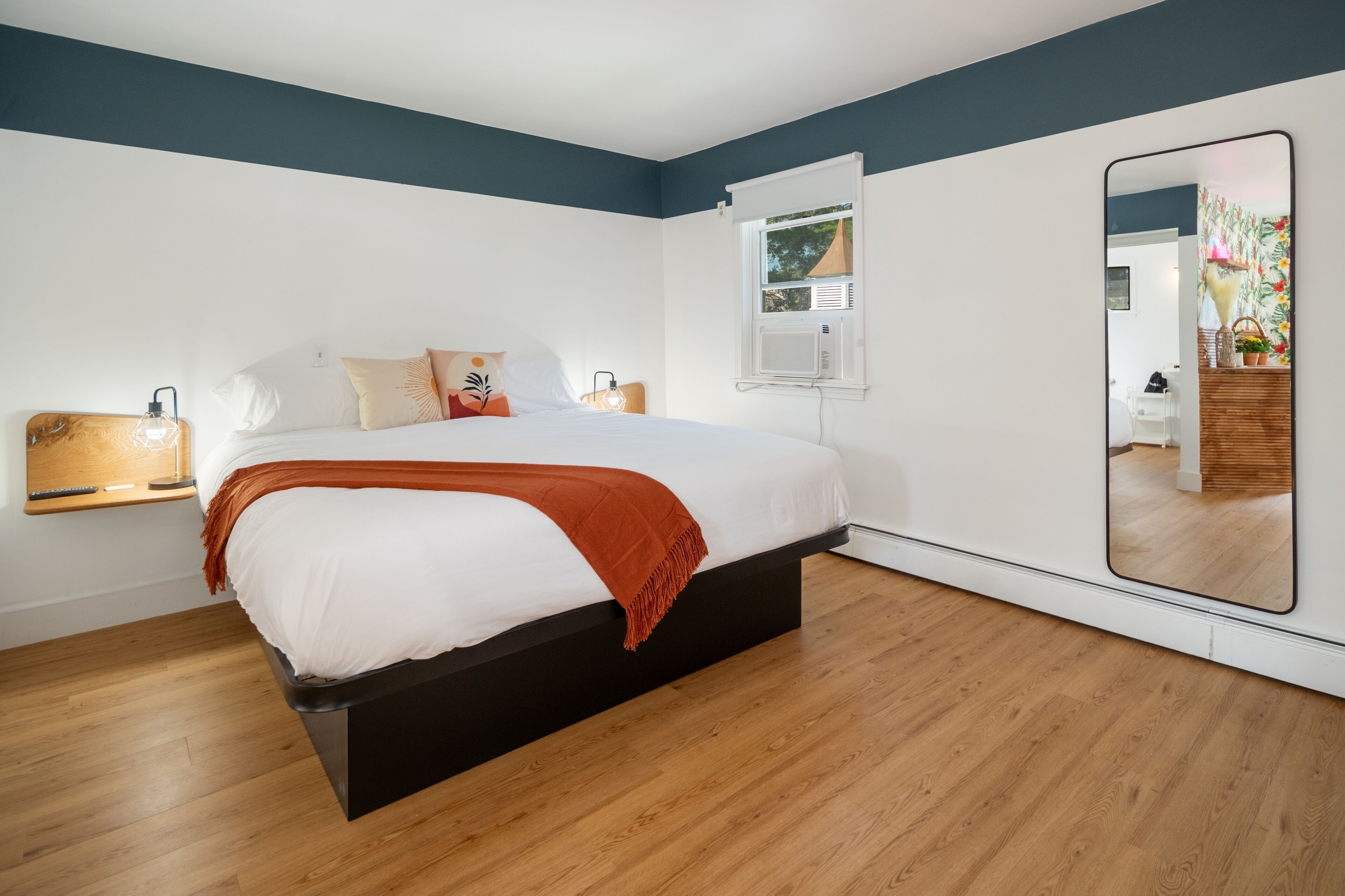 Una camera da letto moderna con un letto, lampade da comodino, uno specchio grande e una finestra con un condizionatore.