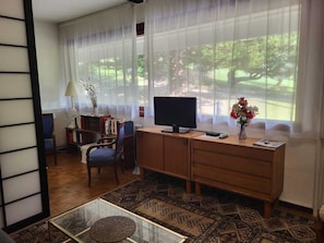 Una habitación con escritorio, silla, televisor, estantería y ventana con vista a vegetación.