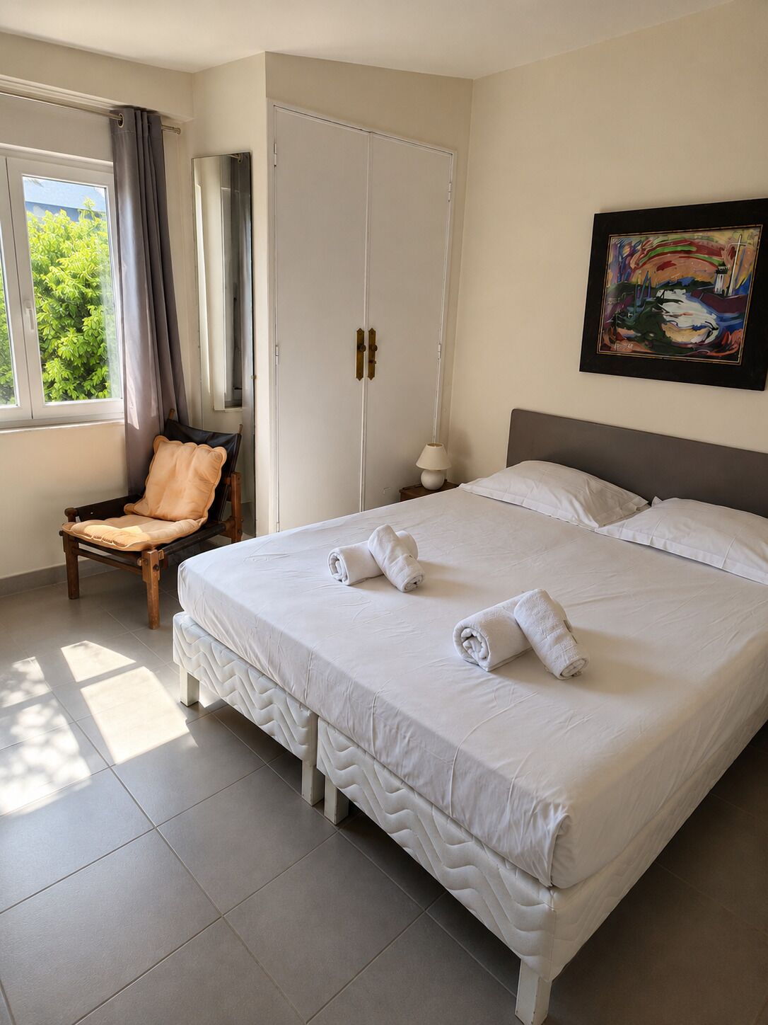 A hotel room with a bed, a chair, a wardrobe, a painting, and a window with curtains.