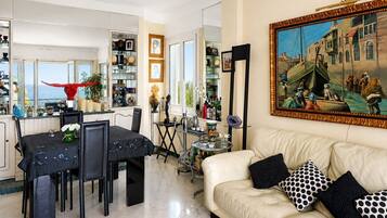 Una sala de estar con un sofá color beige, almohadones estampados en blanco y negro, una zona de comedor con una mesa dispuesta para cuatro personas, y un cuadro grande de una escena histórica en la pared.