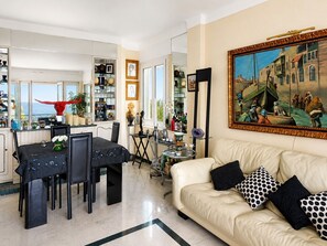 A living room with a beige sofa, black and white patterned pillows, a dining area with a table set for four, and a large painting of a historical scene on the wall. - Sweet Accommodation (Cannes)