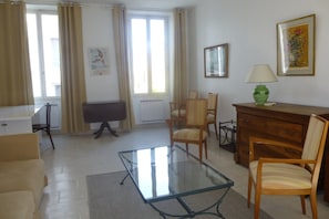 Un salon avec une table basse en verre, un canapé beige, des chaises en bois, un bureau en bois et une lampe.