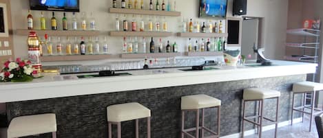 A bar area with a marble floor, multiple liquor bottles on shelves, bar stools, and two TVs mounted on the wall.