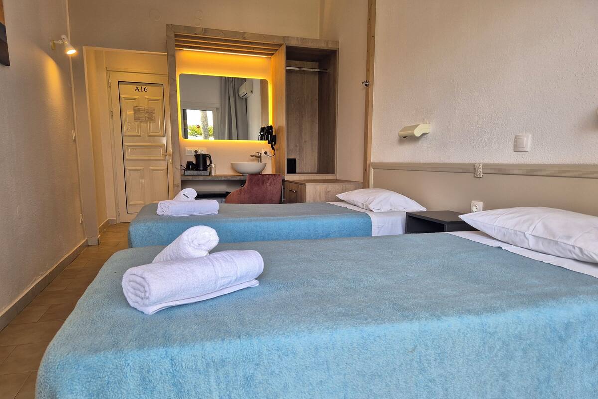 Une chambre d’hôtel avec deux lits, chacun recouvert d’une courtepointe bleue et agrémenté d’oreillers blancs. On y trouve des serviettes sur les lits, une tête de lit en bois, un miroir et une petite table avec une bouilloire et des tasses.