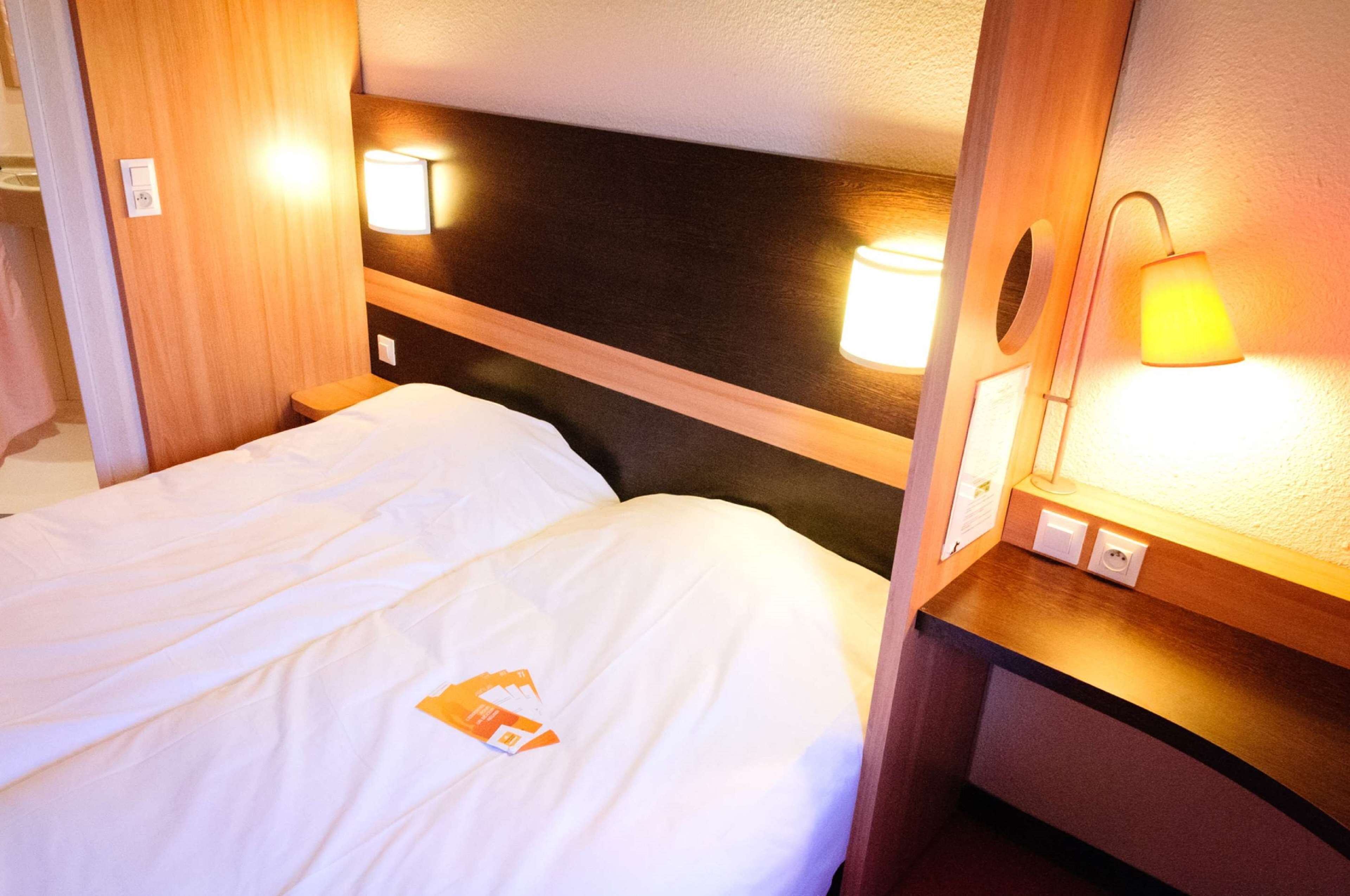 A hotel room with a bed, a wooden headboard, two wall-mounted lamps, and a small table with a lamp.