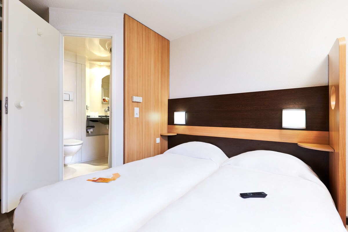 A hotel room with two beds, a wooden headboard, a small bedside table, and a bathroom visible through an open door.