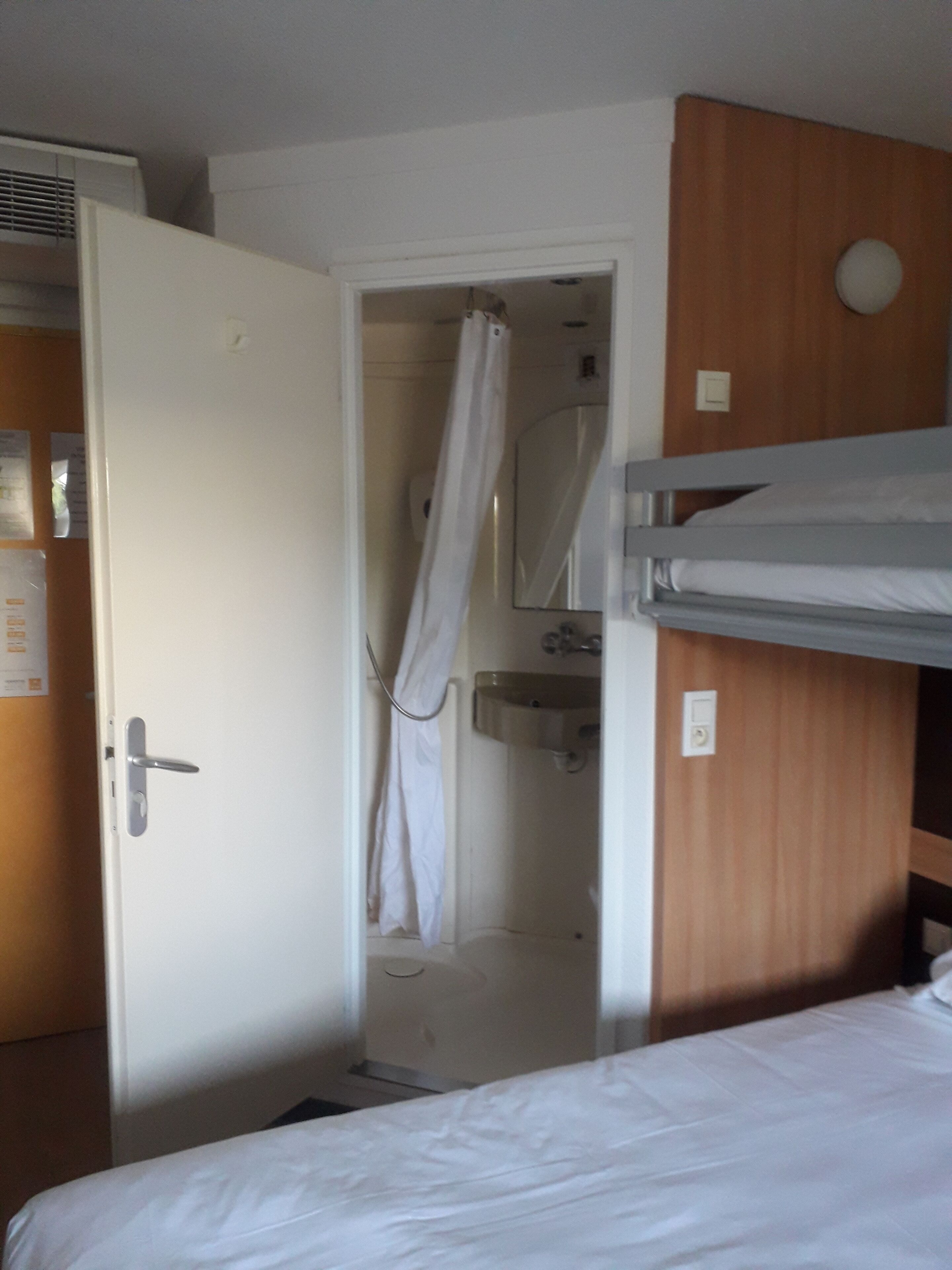 A hotel room with a bunk bed, a shower with a curtain, a sink, and a door with a handle.