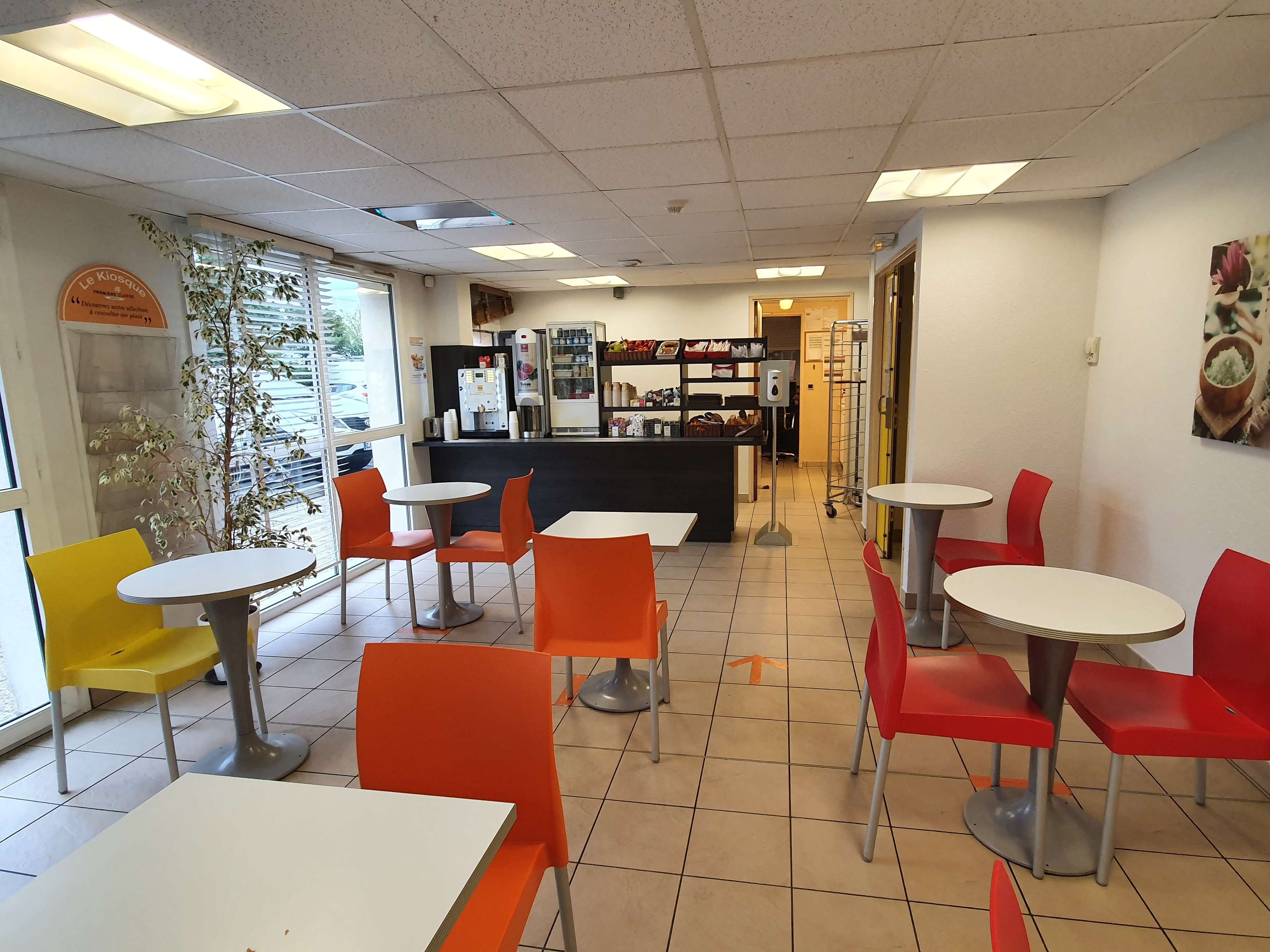 A restaurant interior with multiple tables and chairs, a counter with a beverage dispenser, and a menu board on the wall.