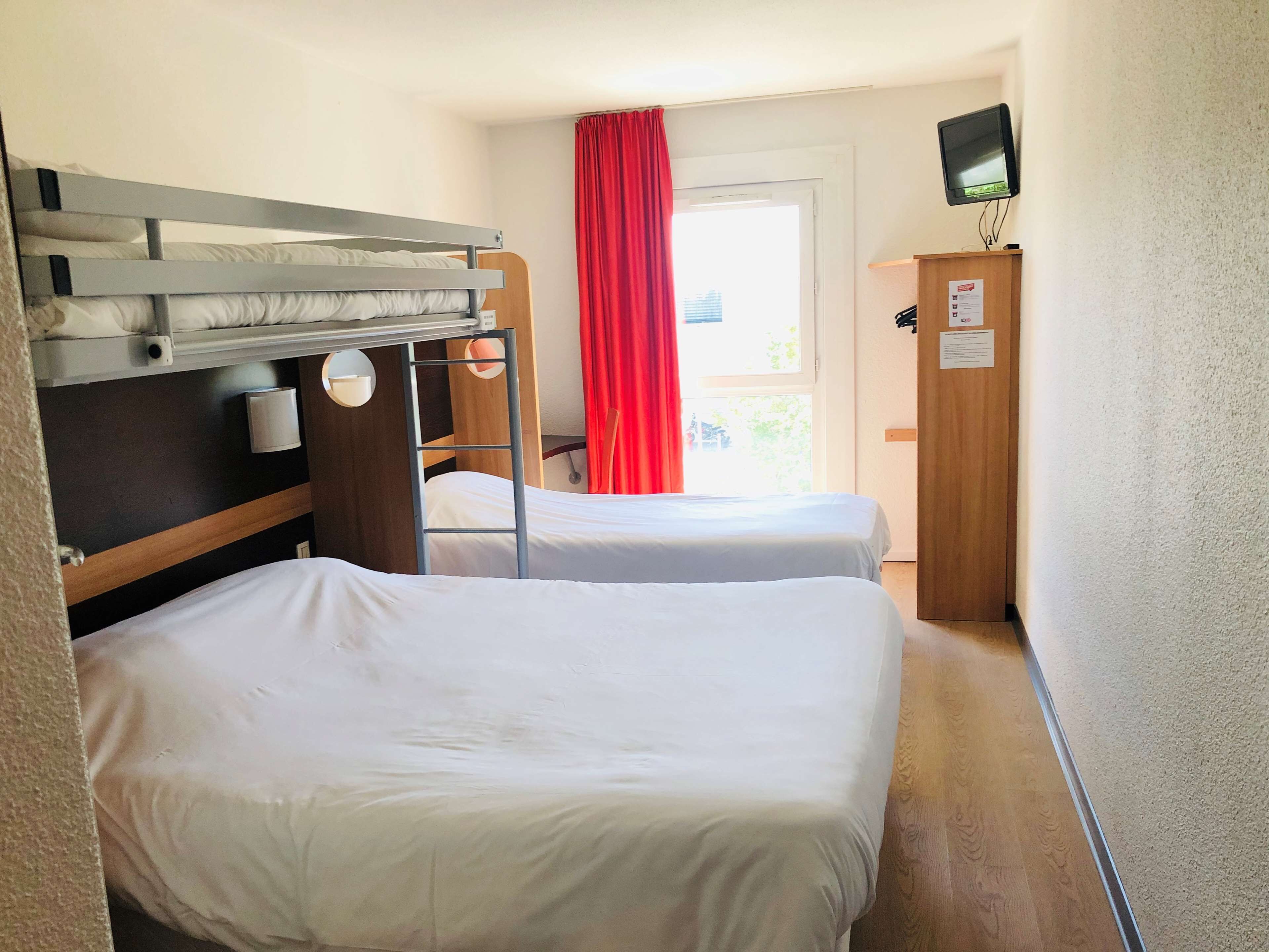 A hotel room with two single beds, a bunk bed, a wooden wardrobe, a television mounted on the wall, and a window with red curtains.