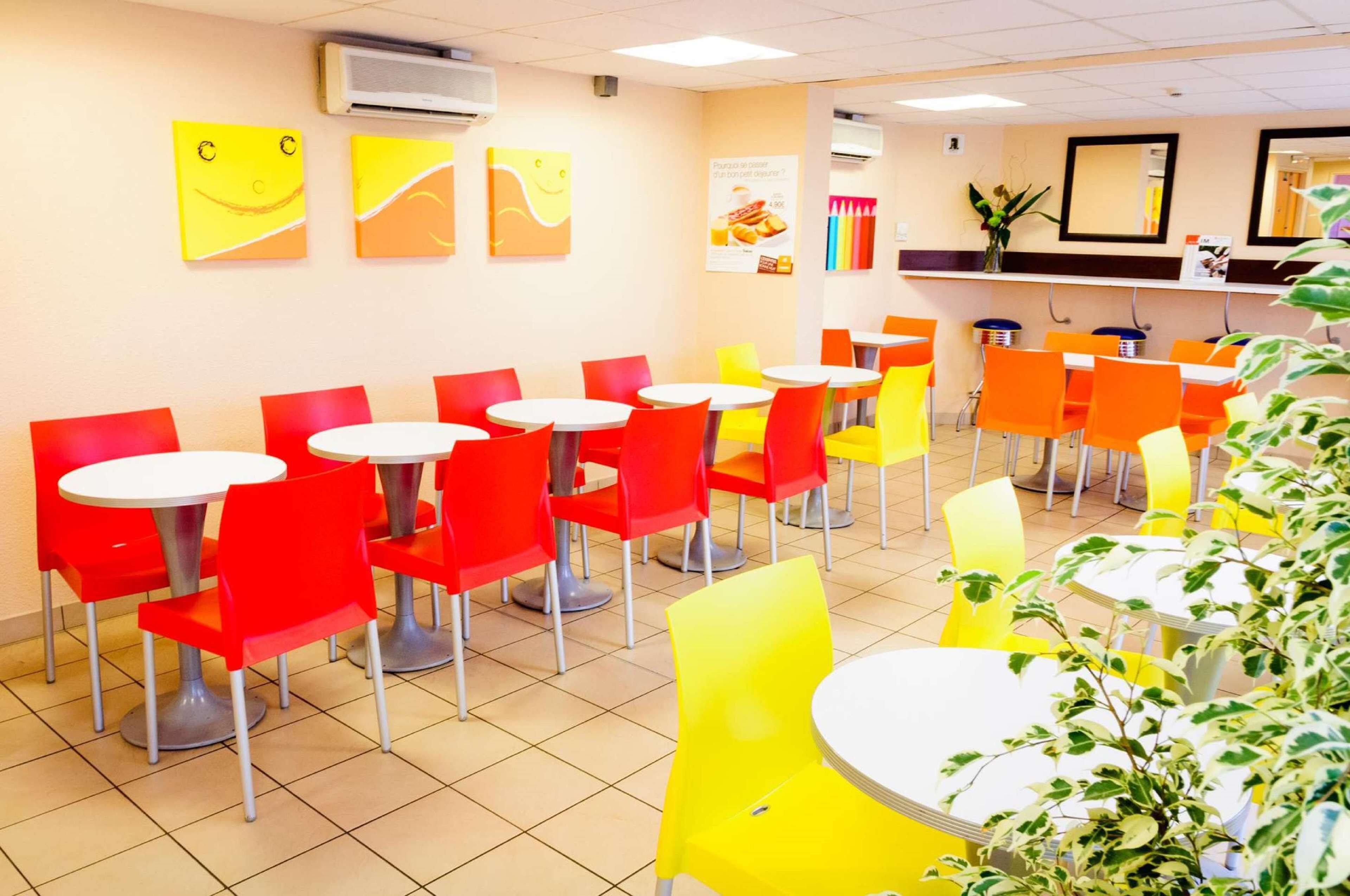 A brightly lit indoor dining area with red and yellow chairs and tables, air conditioning units on the ceiling, and wall decorations.
