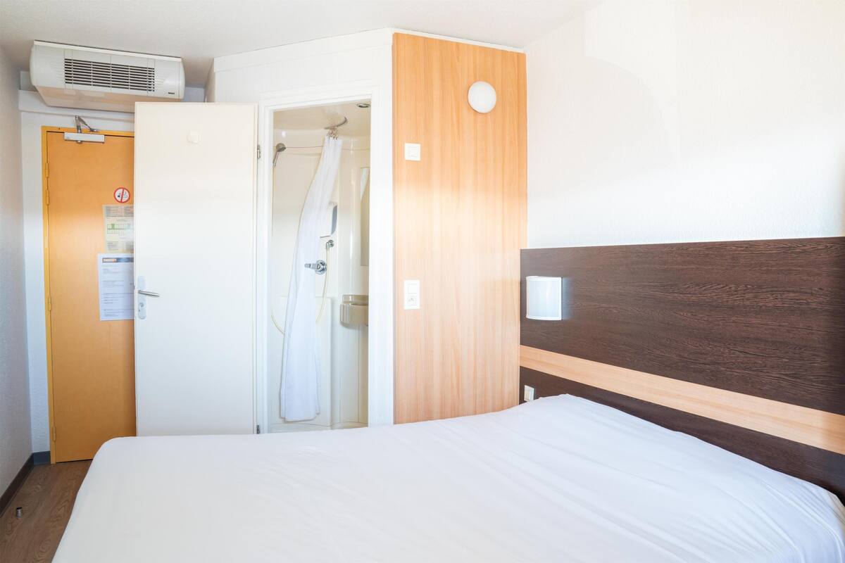 A hotel room with a bed, a wooden headboard, a white door, and a shower area.