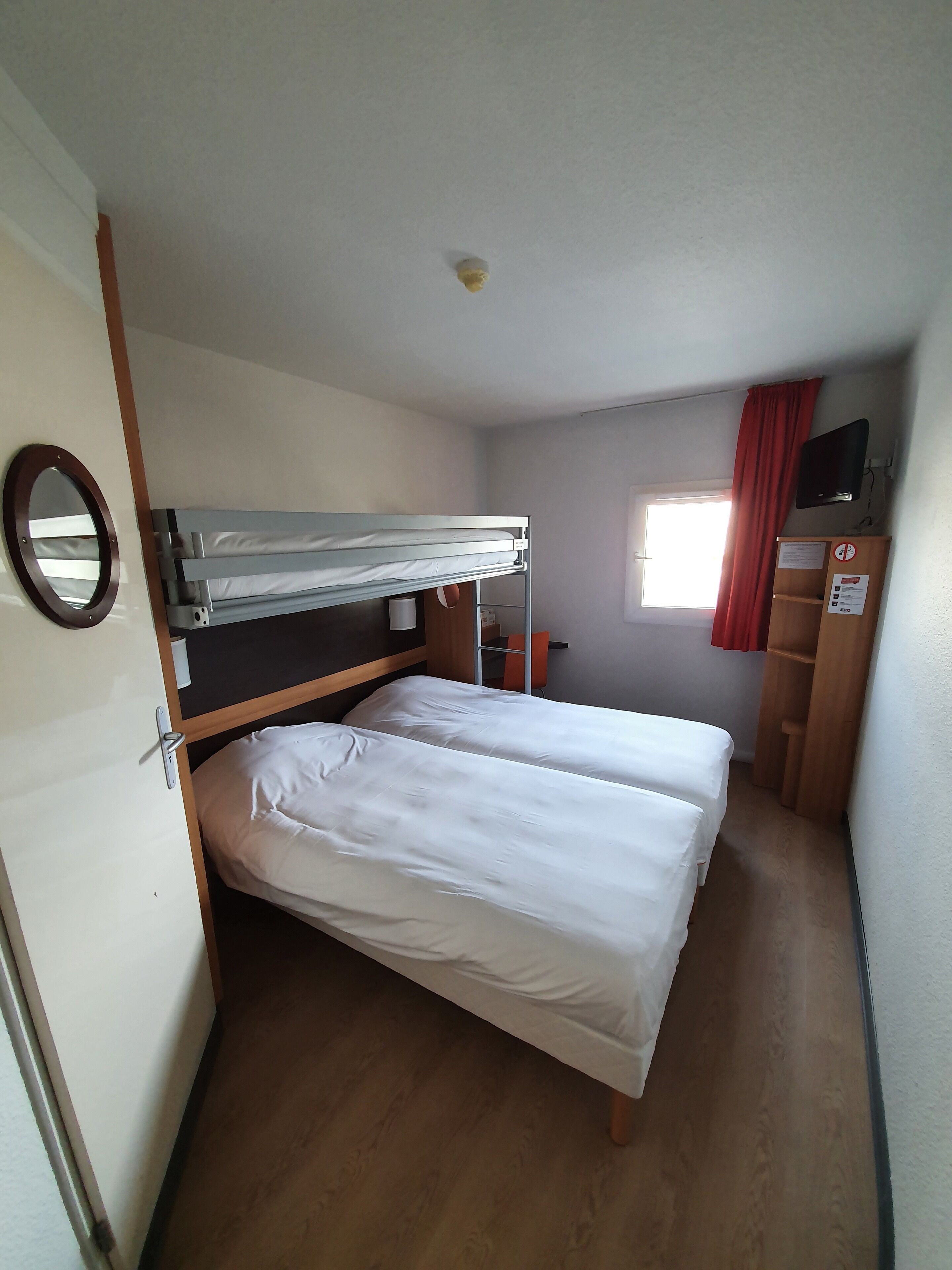Een kleine kamer met een stapelbed, een bureau, een spiegel en een raam met gordijnen.