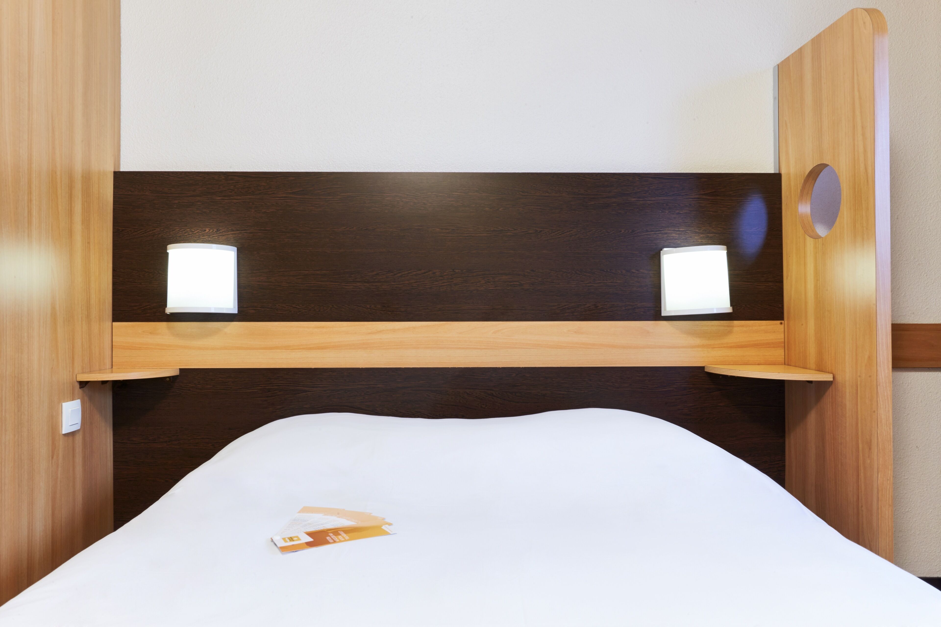 Una cama individual con colchón blanco y cabecero de madera. Hay dos mesitas de noche con lámparas integradas.
