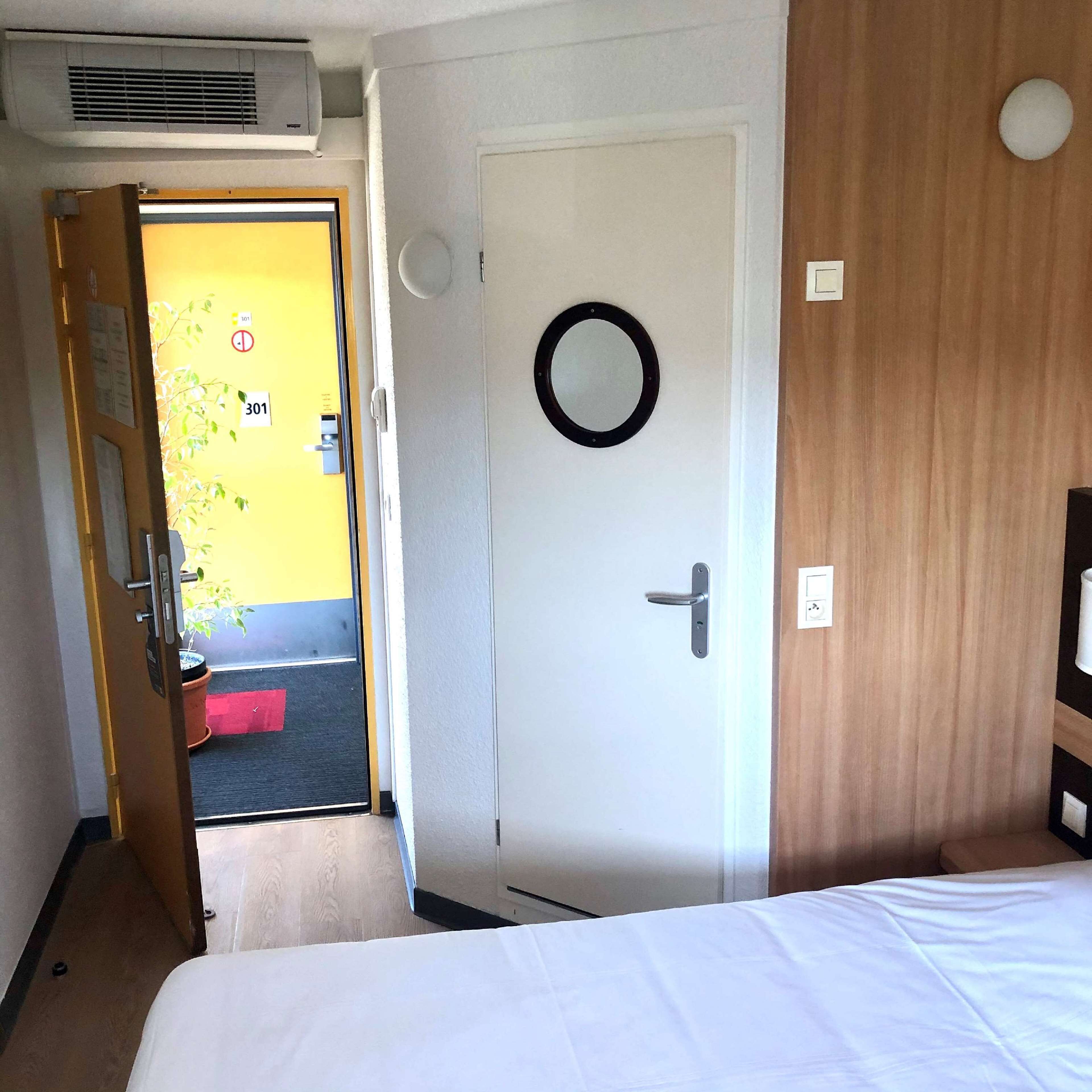 Habitación de hotel con una cama, una puerta blanca con un espejo circular, un armario de madera y una pared amarilla con el número 301.