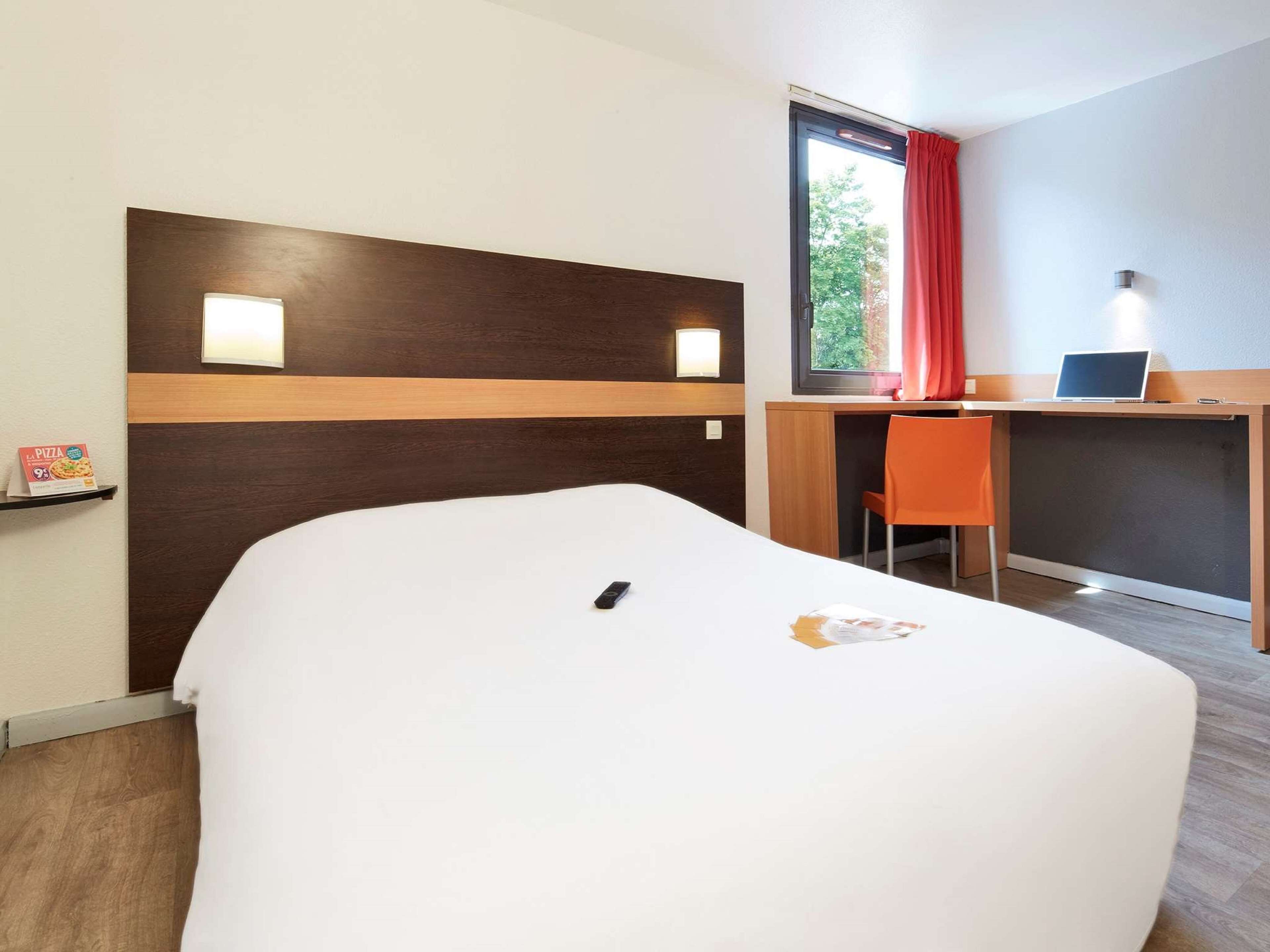 Quarto de hotel com uma cama, uma escrivaninha e uma cadeira. Há uma janela com cortina vermelha, uma prateleira na parede com uma caixa de pizza, e um laptop na escrivaninha.