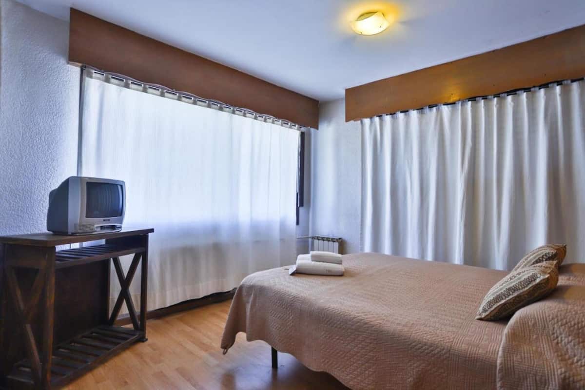 Una habitación de hotel con una cama, una mesita con televisor y cortinas en las ventanas.