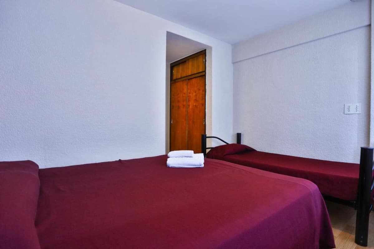 Una habitación con dos camas, cada una con una colcha roja y una toalla blanca sobre una de ellas. Se ve una puerta de madera al fondo.