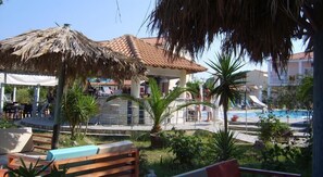 A pool area with lounge chairs, a thatched umbrella, and a building with a tiled roof. - Sweet Dreams Apartments (Corfu)