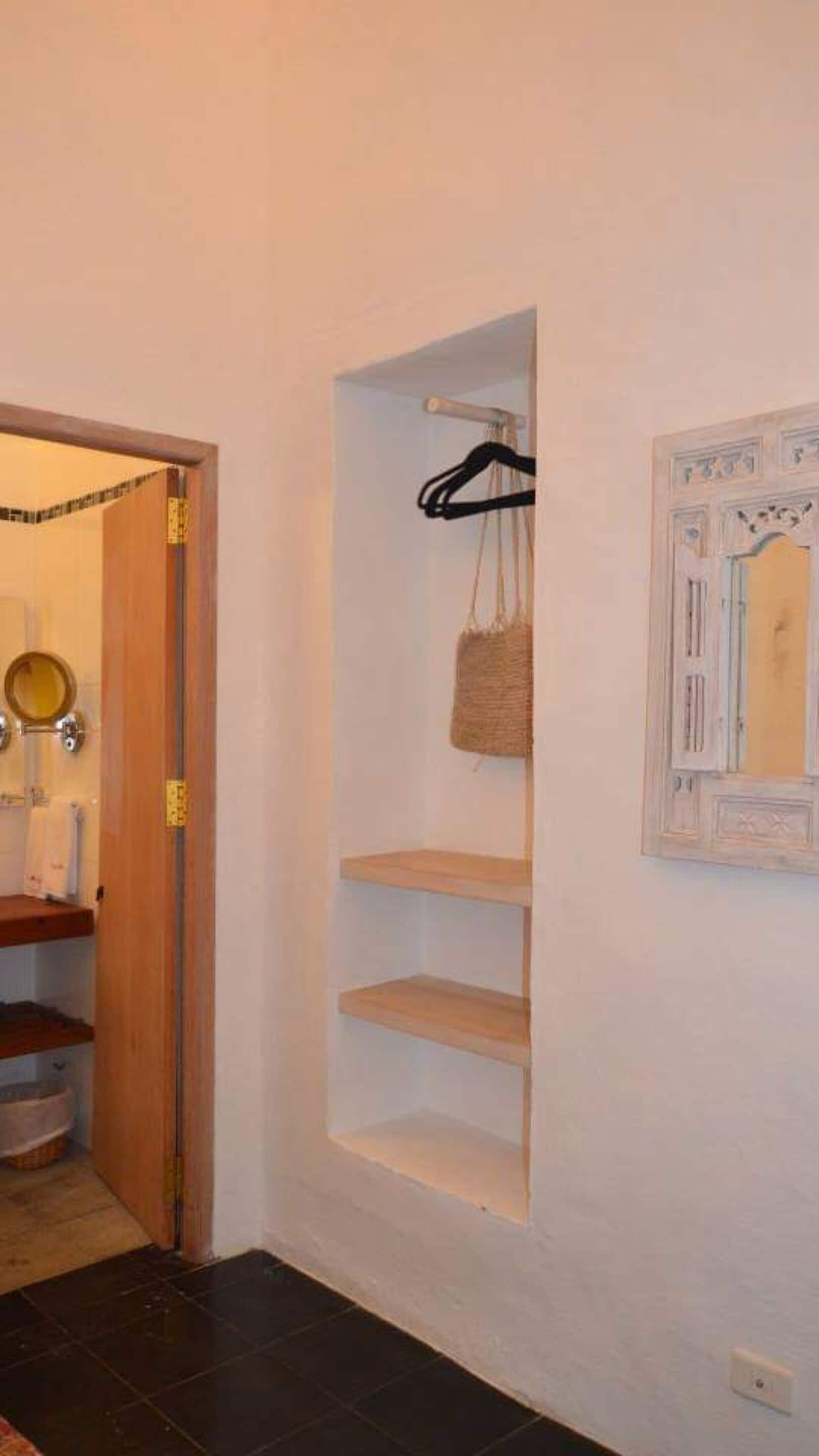A hallway with a wooden door, a mirror, and a wall-mounted shelf with a hanging bag.