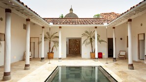 Un patio con piscina central, columnas, palmeras en macetas y una estructura abovedada al fondo.