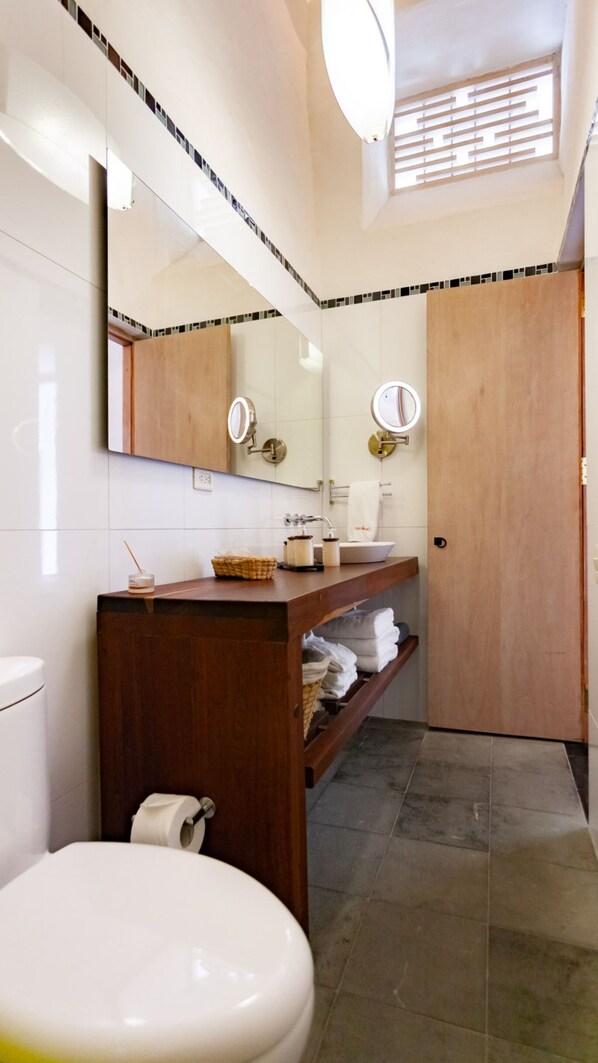 A bathroom with a toilet, sink, and mirror. There is a towel rack and a shelf with towels.