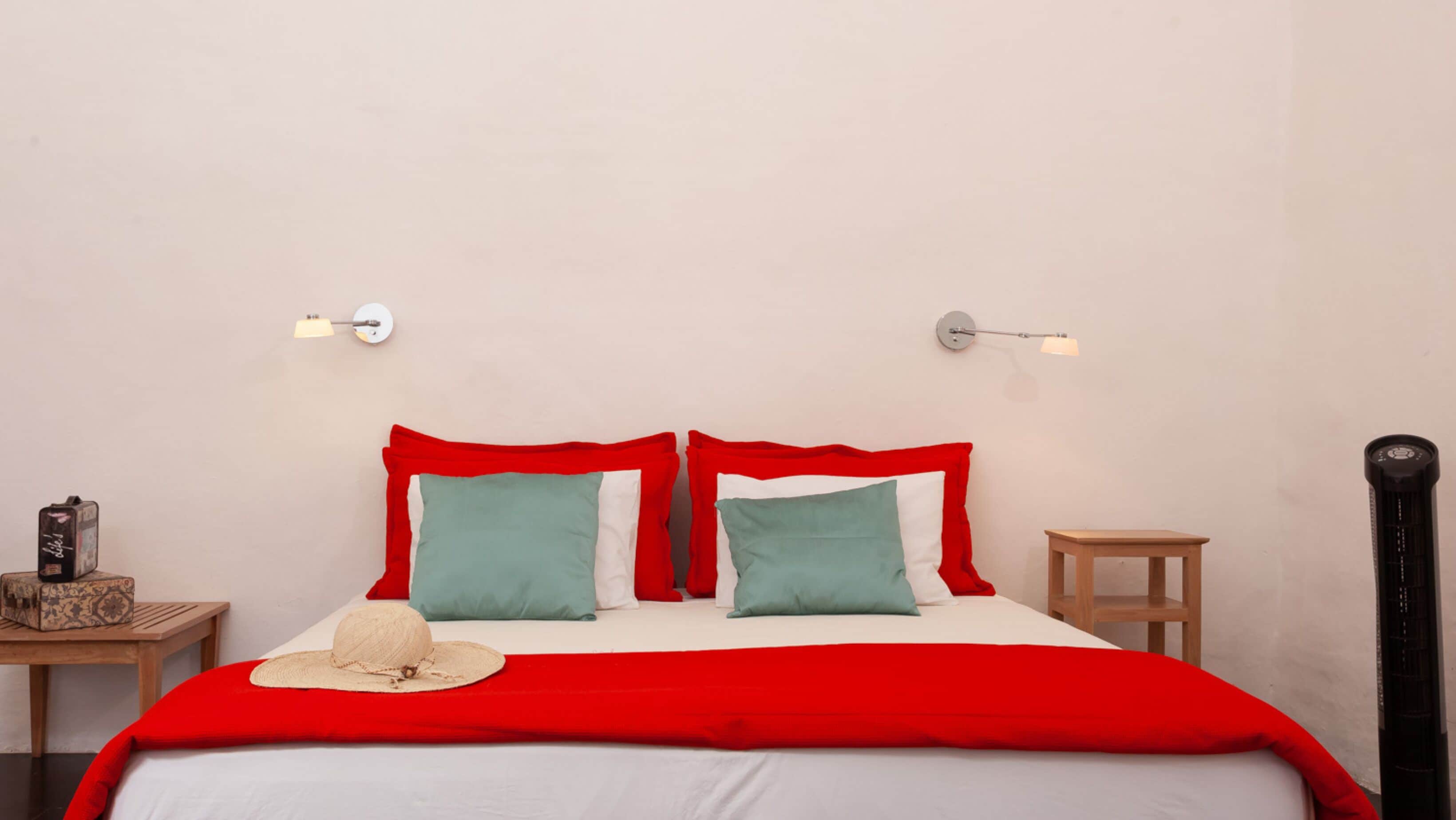 Una cama con ropa de cama roja y blanca, dos almohadas, una mesita de noche de madera, una lámpara y un radiador negro alto.