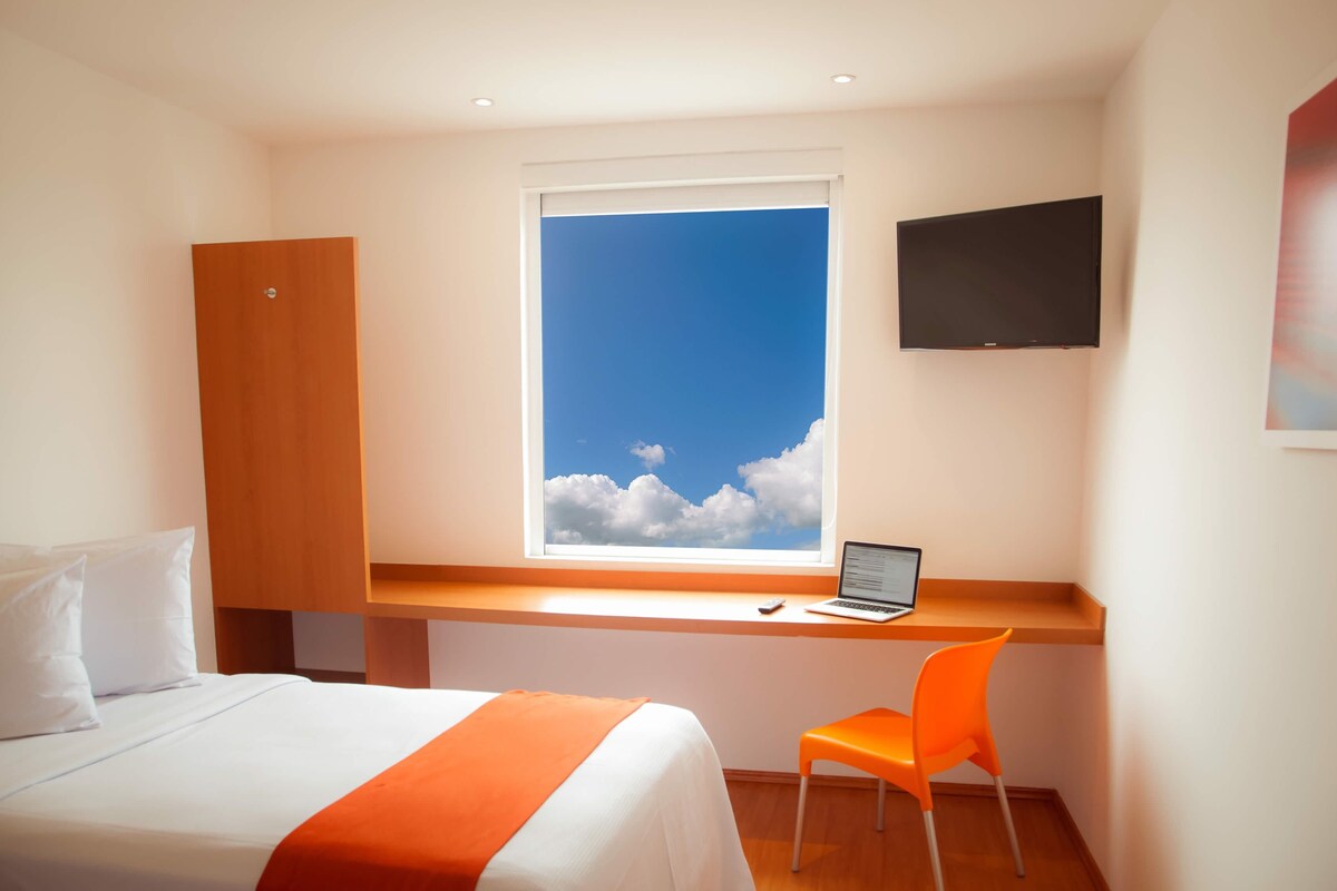 Ett hotellrum med en säng, ett skrivbord med en bärbar dator, en tv och ett fönster med utsikt över en klar himmel.