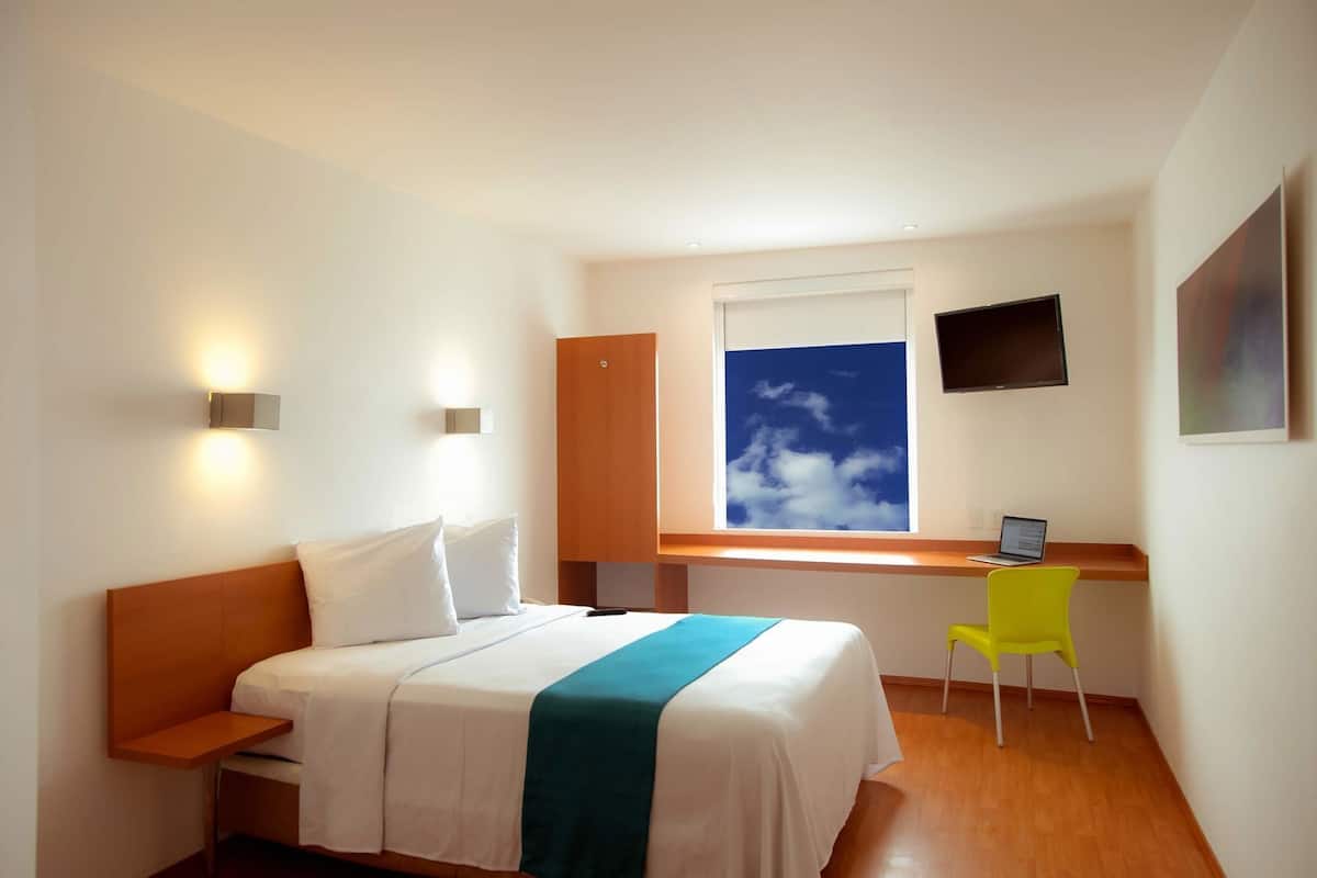 Habitación de hotel con cama, escritorio, silla, televisión y ventana con vista al cielo.