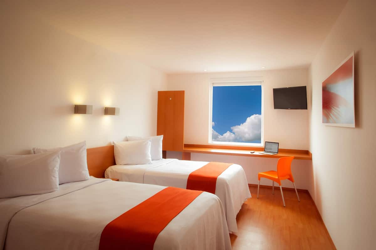 Habitación de hotel con dos camas, un escritorio con una laptop, un televisor y una ventana con vista al cielo.