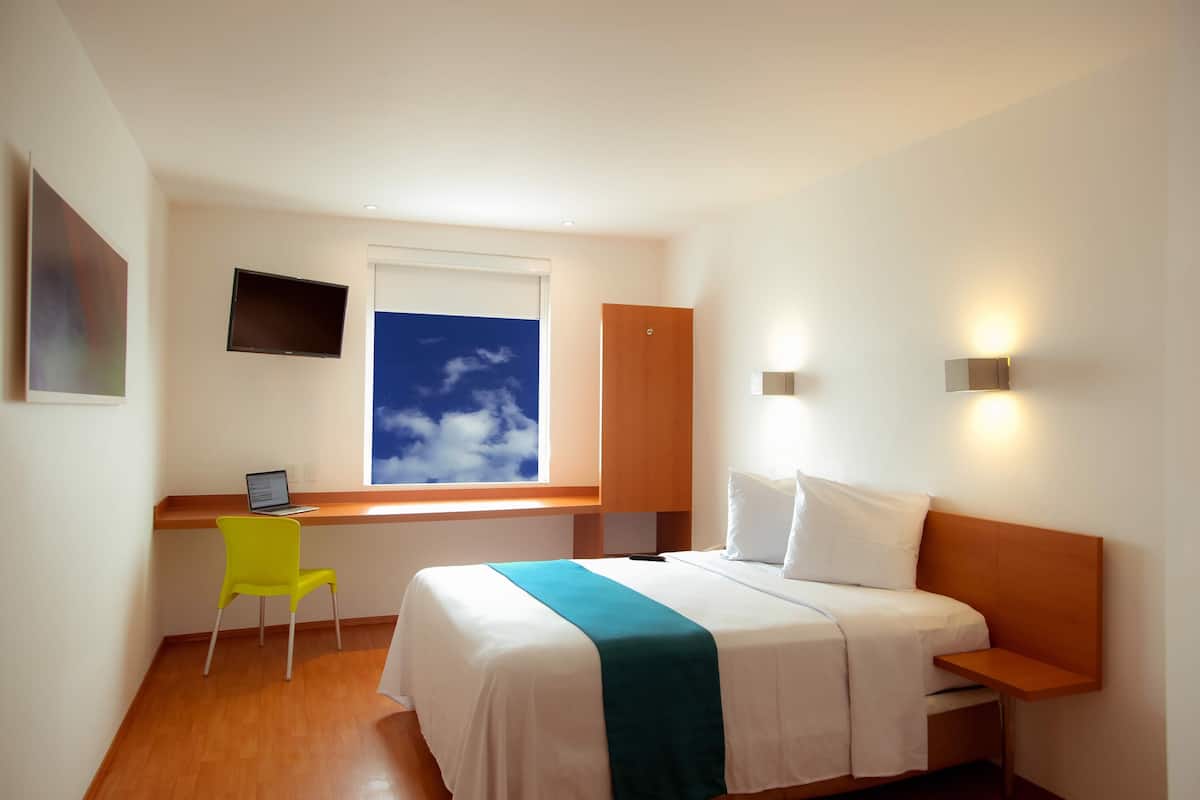 Habitación de hotel con cama, escritorio, silla, televisor y ventana con vistas al cielo.