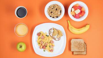 Un plato de desayuno con huevos revueltos, salchicha y tostadas, un plátano, una manzana verde, un vaso de jugo de naranja, un tazón de granola, un tazón de fruta y una taza de café sobre un fondo naranja.