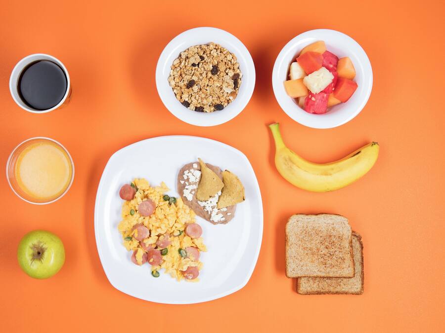 Un plato de desayuno con huevos revueltos, salchicha y tostadas, un plátano, una manzana verde, un vaso de jugo de naranja, un tazón de granola, un tazón de fruta y una taza de café sobre un fondo naranja.