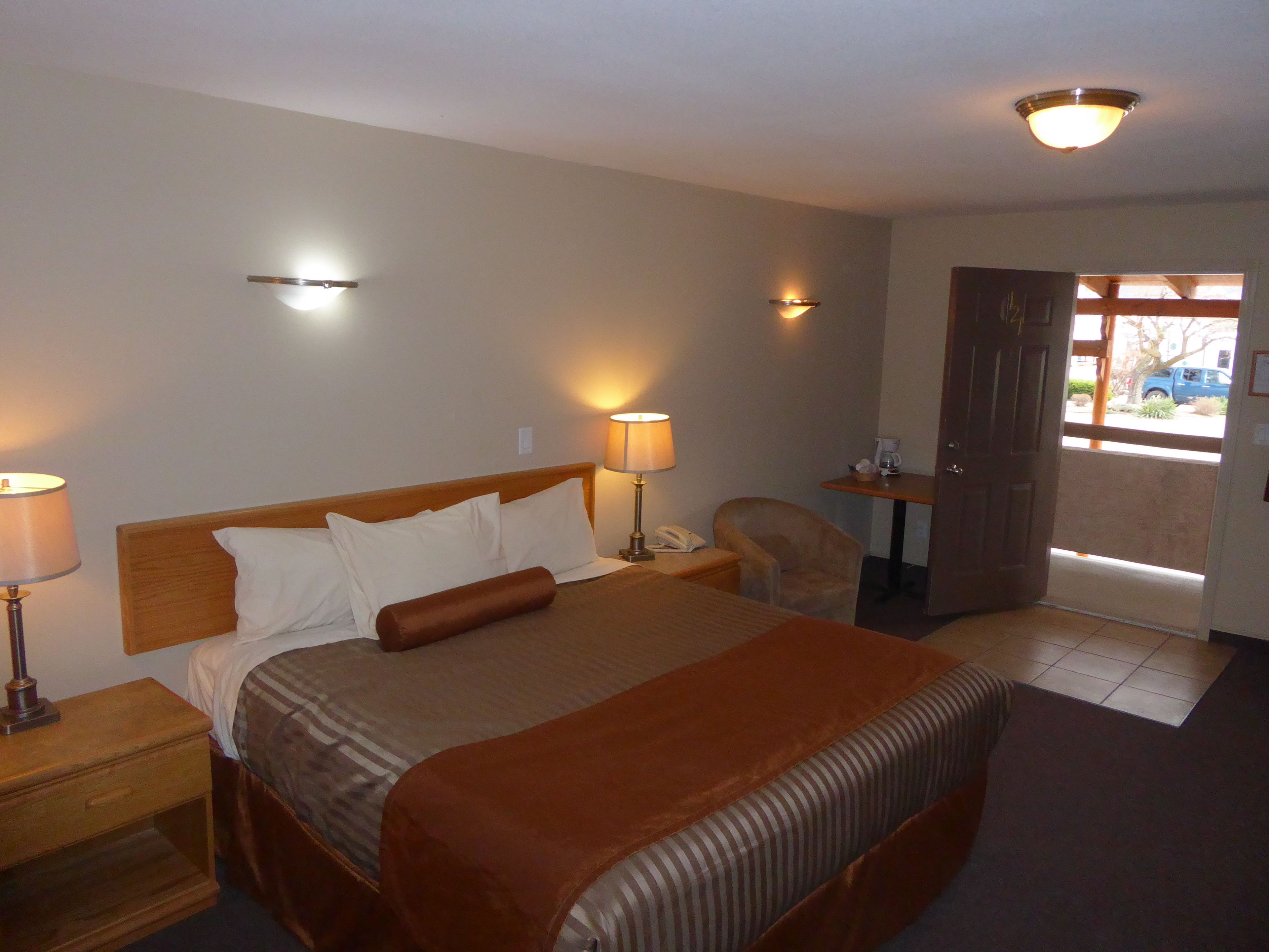 A hotel room with a large bed, bedside lamps, a chair, a small table with a lamp, and a door leading outside.