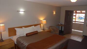 A hotel room with a large bed, bedside lamps, a chair, a small table with a lamp, and a door leading outside.