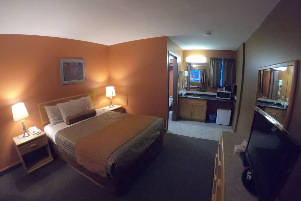 A hotel room with a bed, bedside lamps, a television, a small kitchenette, and a bathroom.