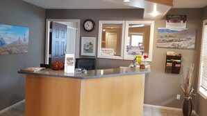 A reception area with a wooden reception desk, a clock, framed pictures, and a window with blinds. - Avalon Inn (Osoyoos)