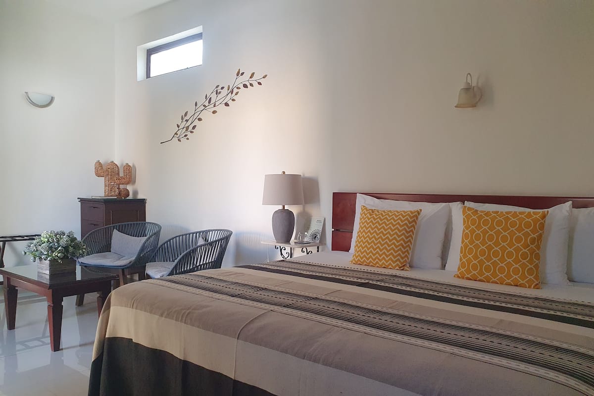 A hotel room with a large bed, a desk with a lamp, a chair, and a small table with a plant.