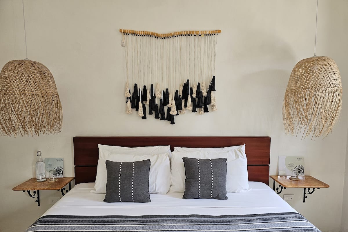 A neatly arranged bedroom with a large bed, two bedside tables, a hanging macrame wall decoration, and two woven pendant lights.