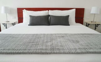 Una cama bien hecha con edredón blanco y dos almohadas, una manta de color gris con estampado y un cabecero rojo.