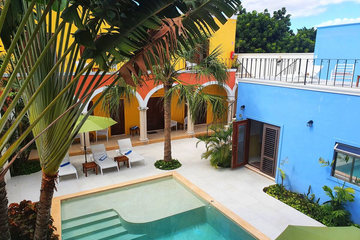 A pool area with lounge chairs and a small pool, surrounded by palm trees and a building with yellow and blue walls.